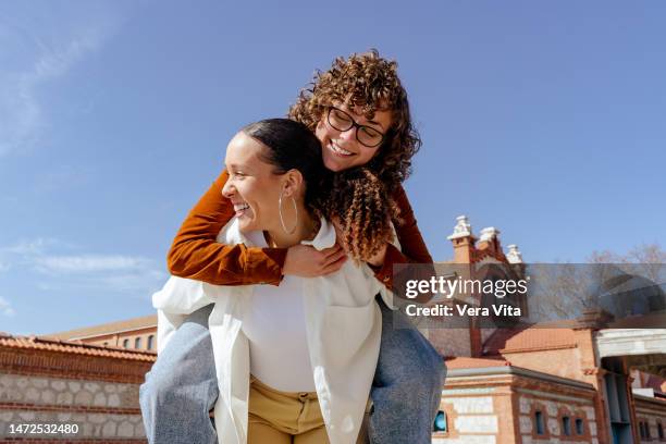 funny portrait of caucasian and afro women friends piggybacking with happiness outdoors - afro jokes stock-fotos und bilder