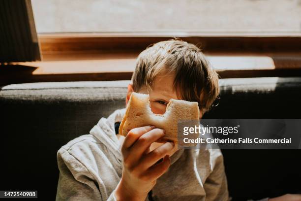 a young boy playfully peers through a bite he has just taken out of his sandwich - eating bread stockfoto's en -beelden