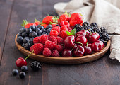 Fresh organic summer berries mix in round wooden tray on dark wooden table background. Raspberries, strawberries, blueberries, blackberries and cherries with linen kitchen towel. Space for text