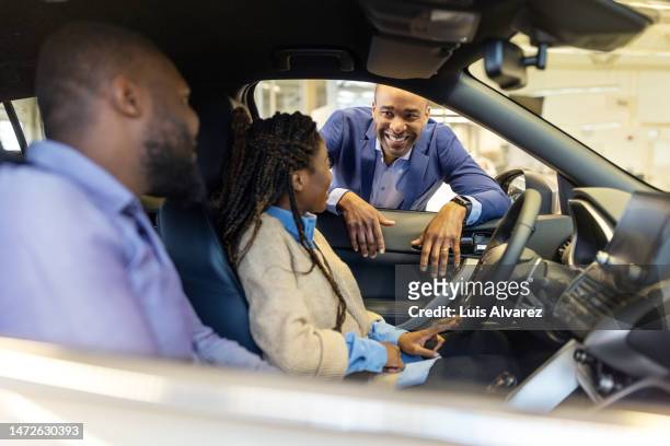salesman explaining the new features to african couple sitting inside the car at showroom - car dealership test drive stock pictures, royalty-free photos & images