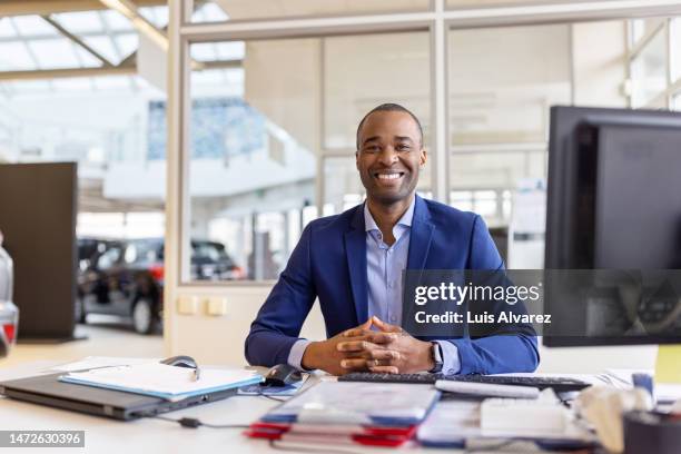 portrait of a confident car dealer sitting at his office desk and smiling - friendly salesman fotografías e imágenes de stock