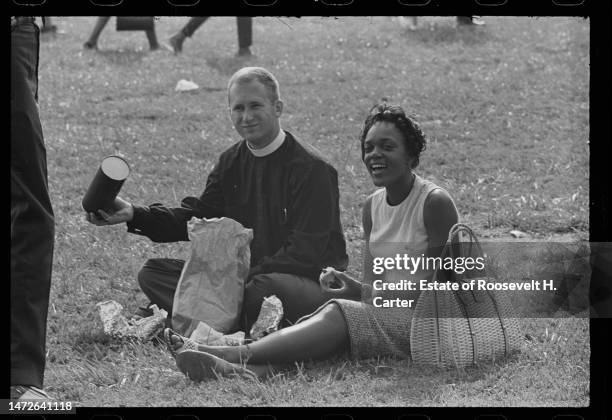 An unidentified man in a clerical collar and Patricia Ann Isbell smile as they sit on the grass and eat a meal during the March on Washington for...