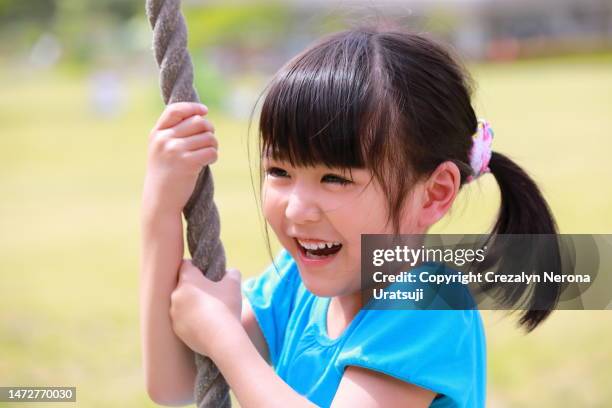 cute little girl in a blue one piece dress having fun at the at the public park playground hanging on a rope swing in close up - 子供のみ ストックフォトと画像