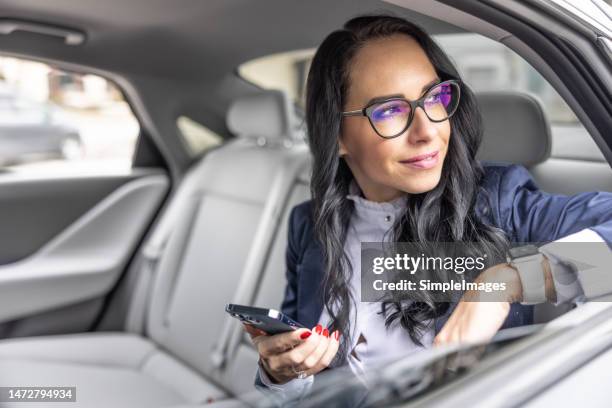 female executive wearing glasses looks outside the window of a luxury car driven through the city. - chauffeur stock pictures, royalty-free photos & images