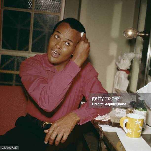 American singer, actor and civil rights activist Harry Belafonte wearing a pink shirt mops his brow as he casts his eyes upwards, sitting in his...