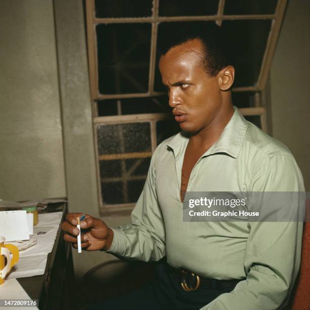 American singer, actor and civil rights activist Harry Belafonte wearing a pale green shirt, with an unlit cigarette in his hand, sitting in his...