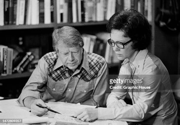 American politician Jimmy Carter, wearing a shirt with plaid patches on the shoulders, and his wife, Rosalynn Carter, reading a document on the table...