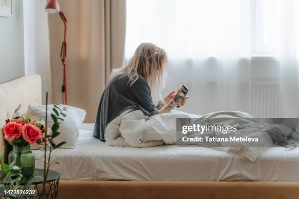 blond long haired female sit in white bed and using phone against the light window view near red flowers on bedside table in bedroom - waking up stock pictures, royalty-free photos & images