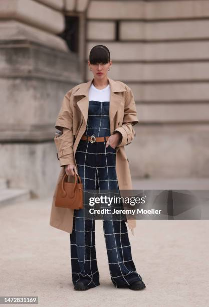 Katya Tolstova is seen wearing a beige trench coat, a blue patterned denim jumpsuit, a brown leather belt and a matching brown leather bag and a...