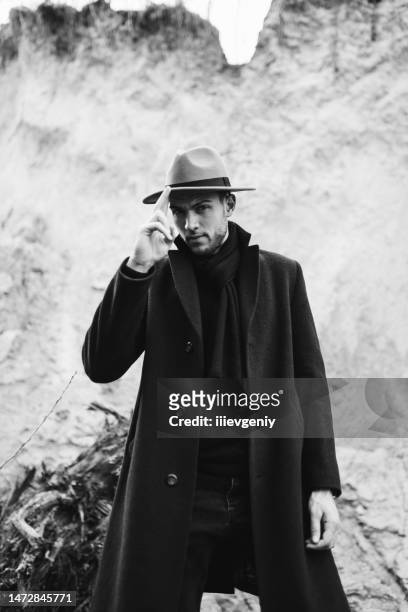 männliches porträt. brünetter mann mit hut und mantel im wald. schwarz-weiß-fotografie. noir detektiv - male model stock-fotos und bilder