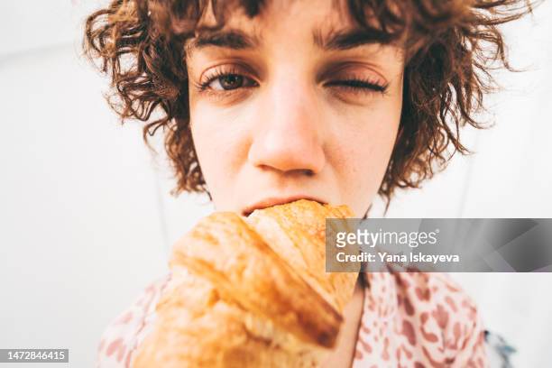 a funny portrait of a cute french young woman eating a giant croissant - vreten stockfoto's en -beelden
