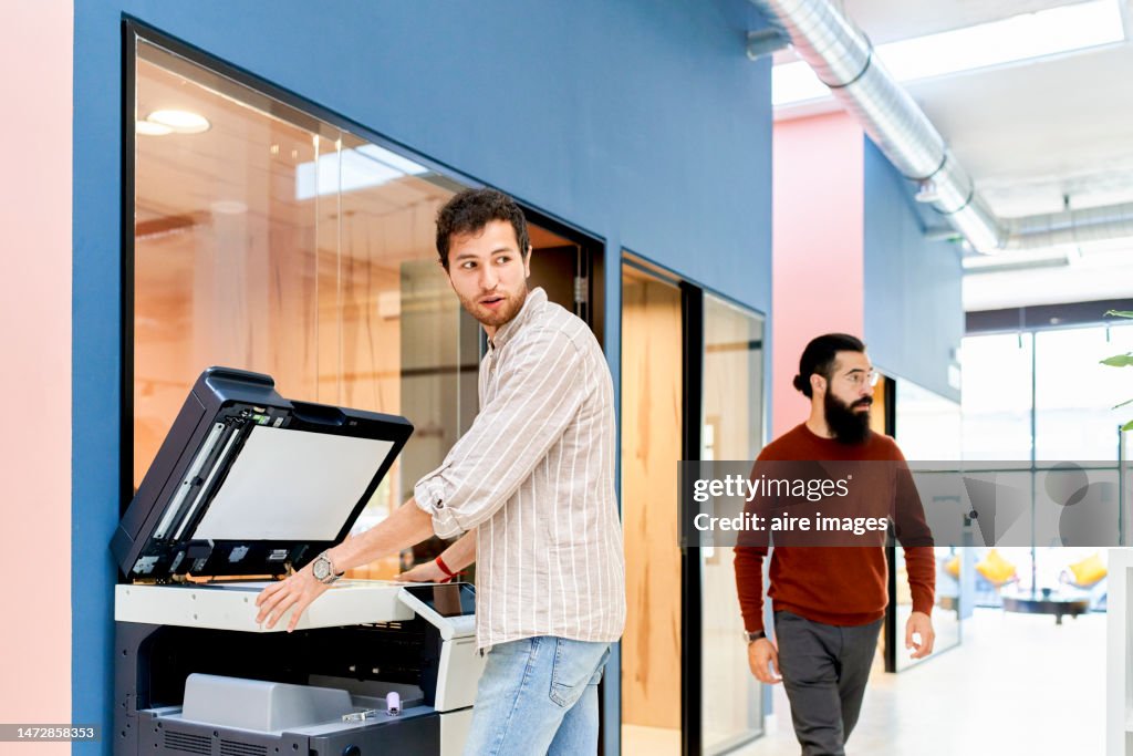 Young man standing in front of the printer talking with a co-worker while scanning documents