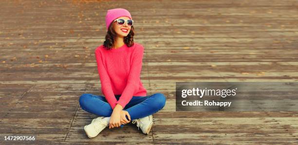 Portrait of stylish smiling young woman wearing knitted sweater and pink hat sitting on wooden floor in the city park portrait stylish smiling young woman wearing