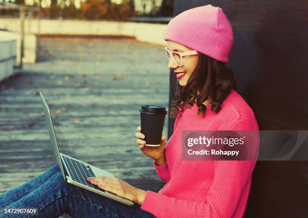 Portrait of stylish happy modern young woman drinking coffee working with laptop wearing a colorful clothes sitting in the city park portrait stylish happy modern young woman