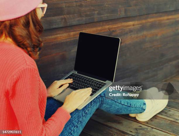Portrait of stylish modern young woman working with laptop wearing a colorful clothes sitting on wooden floor in the city park, black blank screen copy space portrait stylish modern young woman working