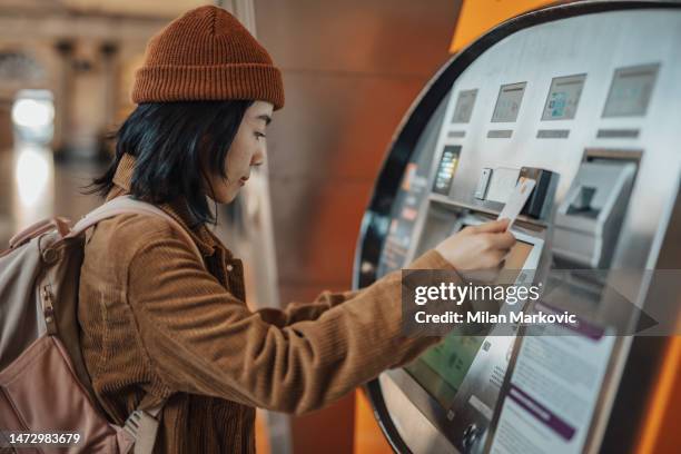 a beautiful young chinese woman uses an atm at a metro station in barcelona, withdraws money to go shopping - transporte público imagens e fotografias de stock