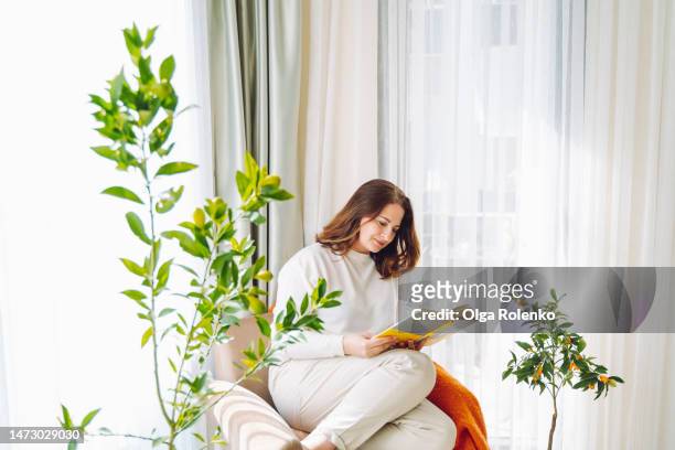 brunette woman is into a book novel, reading, curled up in an armchair with a blanket in light living room - curled up reading book stock pictures, royalty-free photos & images