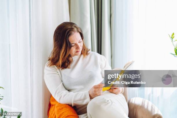 passionate mature woman reading a book in an armchair, near the light window - curled up reading book stock pictures, royalty-free photos & images