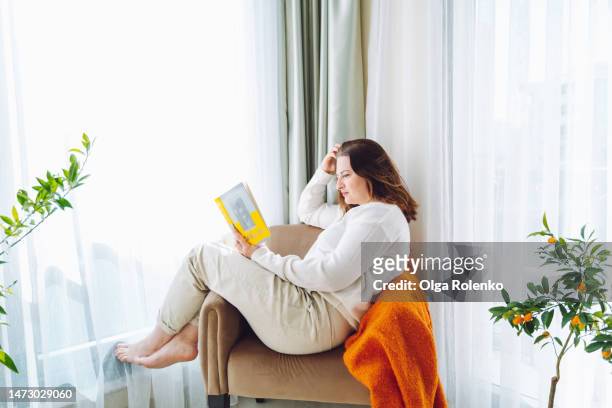 imagination and fantasy with books. barefoot woman reading a book, sitting sideways in an armchair near radiator heater by the window - curled up reading book stock pictures, royalty-free photos & images