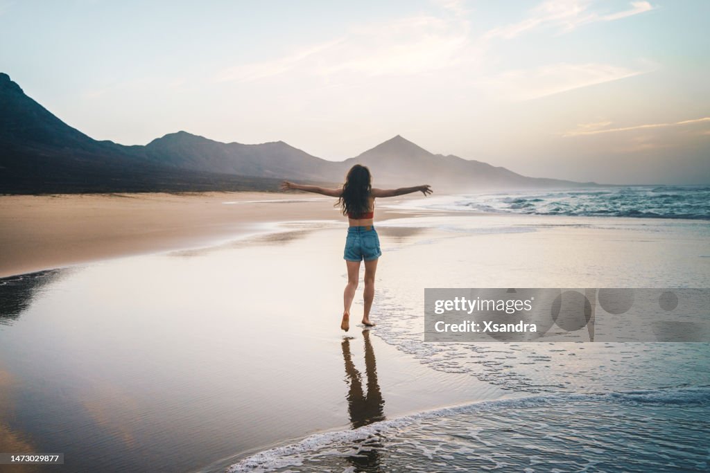 Junge Frau, die bei Sonnenuntergang alleine am Strand spazieren geht - Freiheits- und Glückskonzept