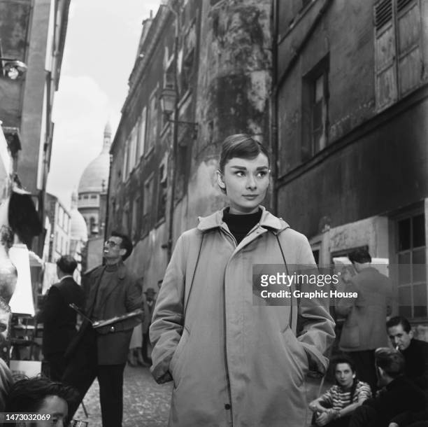 British actress Audrey Hepburn , her hands in the pockets of her raincoat, on a busy street, the dome of Sacre-Coeur visible in the background,...