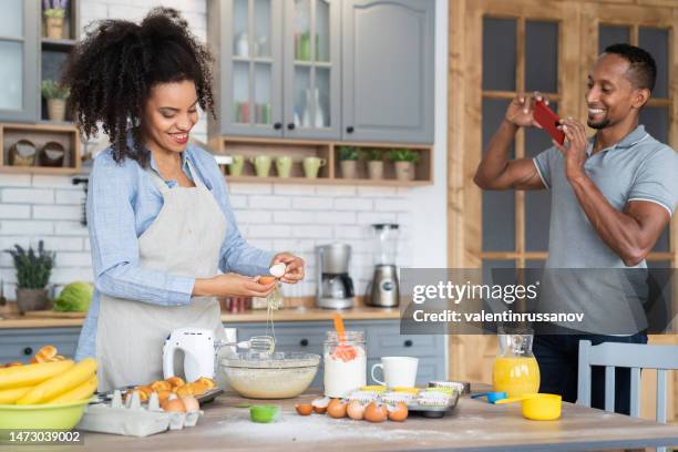 couple cooking together, having fun time in the kitchen. multiracial young woman mixing eggs in a bowl, while her boyfriend taking picture of her with his mobile phone, shooting home video or vlogging - egg icon stock pictures, royalty-free photos & images