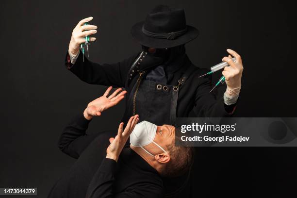 a frightened man dodges away from the plague doctor with syringes. isolated on a black background. covid-19, epidemic and pandemic concept. - bubonic plague mask stock pictures, royalty-free photos & images