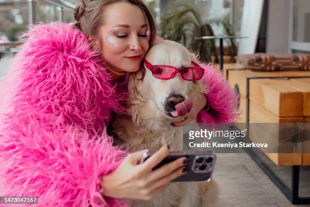 young beautiful european woman in a pink faux fur coat takes a selfie on the phone with a white golden retriever dog in pink glasses - hairy women imagens e fotografias de stock
