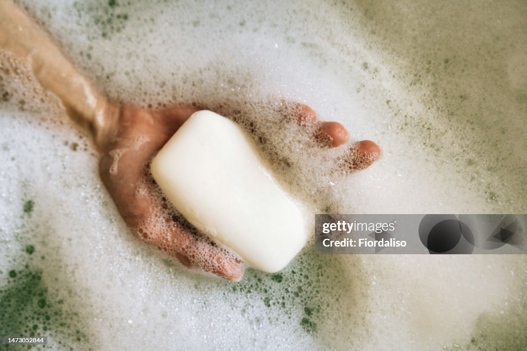 White toilet soap in female hands against the background of a fragrant foam bath. Natural beauty, daily skincare routine. Moisturizing, cleansing