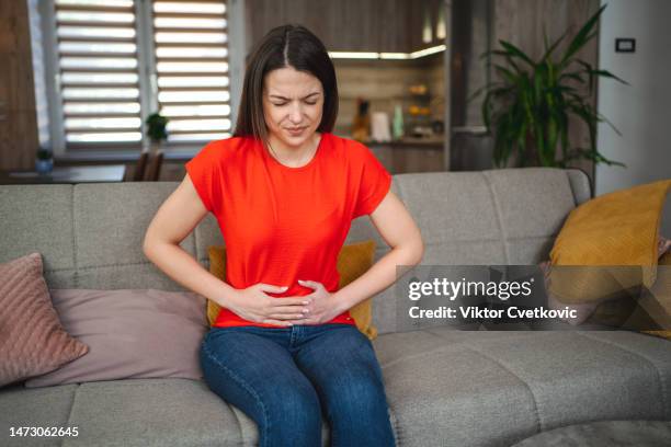 young woman sitting on a sofa experiencing stomach pain - irritable bowel syndrome stock pictures, royalty-free photos & images