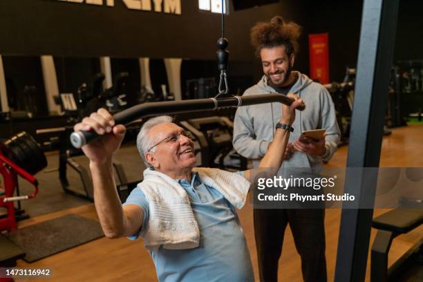 porträt eines älteren mannes mit fitnesstrainer, der kraftgerät im fitnessstudio zieht - rückentraining stock-fotos und bilder