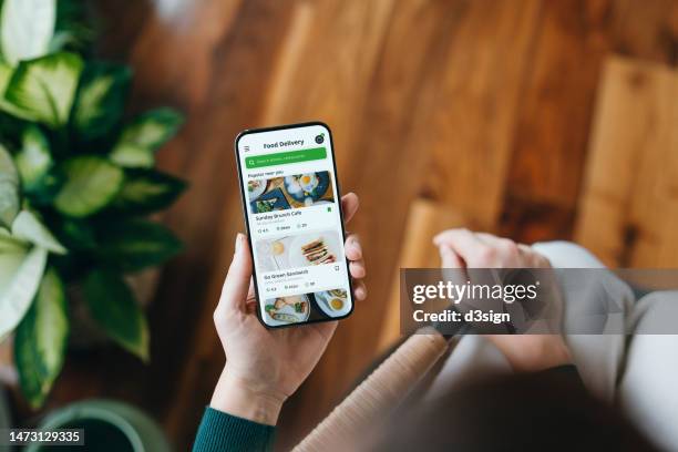 overhead view of young woman's hand using meal delivery service and ordering food online with mobile app on smartphone at home, next to green potted plants. food delivery mobile app. lifestyle and technology - order takeout stock pictures, royalty-free photos & images