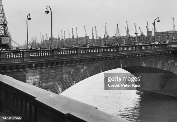 Pedestrians cross old London Bridge [before it's demolition and sale in 1968], cranes and warehouses in the Pool of London line the riverbank in the...