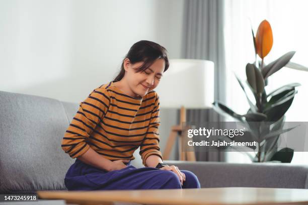 mujer asiática que siente dolor de estómago sentada en el sofá de la sala de estar de su casa. - apendicitis fotografías e imágenes de stock