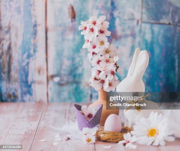 nature morte de pâques d’un œuf dans un nid d’or, d’un lapin de pâques blanc et d’une branche de fleurs d’amandier et d’une grande marguerite sur une table en bois sur fond vintage bleu avec un espace de copie - bunny eggs photos et images de collection