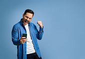 Handsome mid adult man dressed in denim shirt screaming and cheerfully pumping fist while reading good news over smart phone on blue background