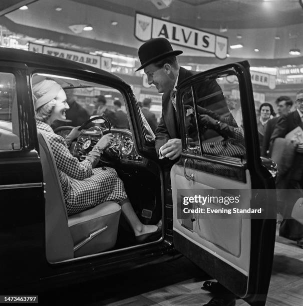 Woman seated at the driver's seat of a new Humber car, smiles at her husband leaning over the open door in a display at the British International...