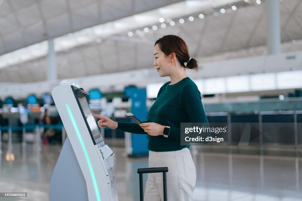 Young Asian woman with suitcase checking-in at the airport using an automated self service check-in kiosk. Ready for a trip. Business travel. Travel and vacation concept