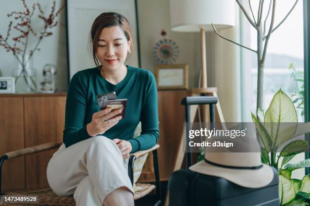 beautiful smiling young asian woman sitting on armchair, holding passport and using smartphone at home, with suitcase and straw hat by her side. ready to go on vacation. business travel. travel and vacation concept - booking hotel foto e immagini stock