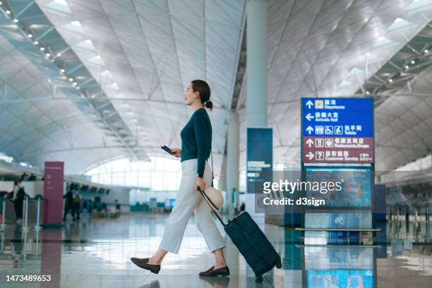 side profile of young asian woman holding passport and boarding pass on hand, walking with suitcase in the airport. young female traveller with sun hat and luggage in airport. ready to go on vacation. travel and vacation concept - card board stockfoto's en -beelden