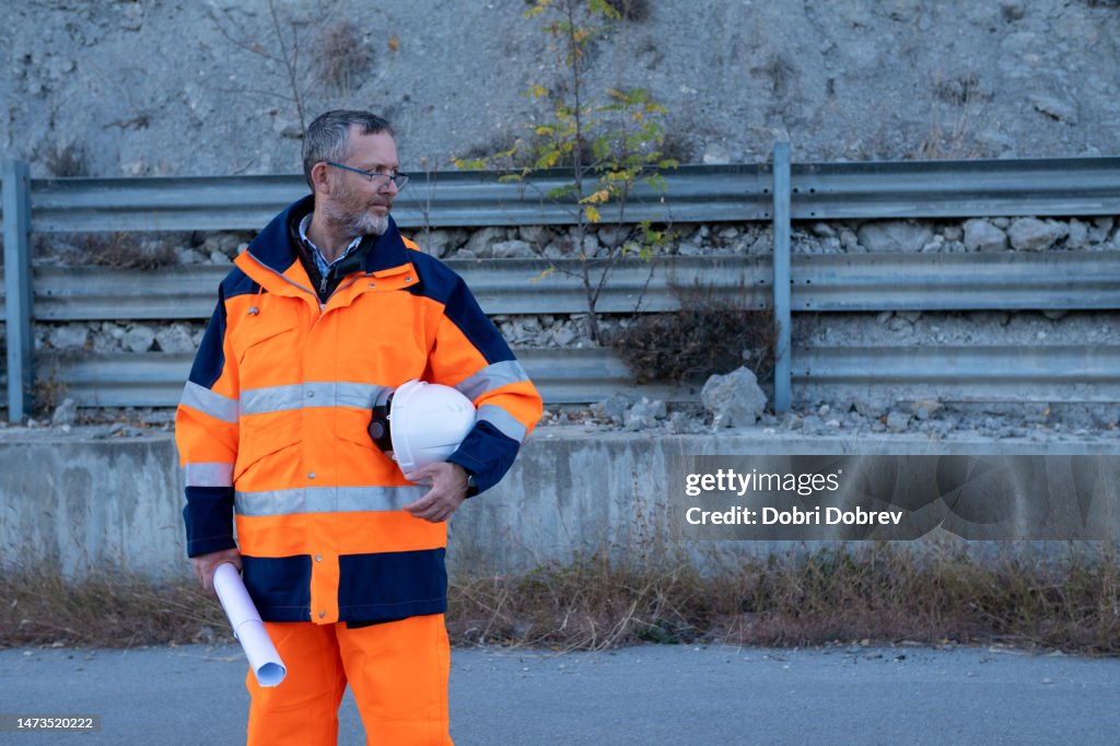 Engineer in white helmet, orange-blue safety reflective suit, and blueprints.