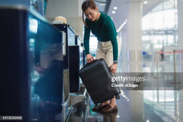 young asian female traveller doing luggage check-in at the airline counter in the airport, putting her suitcase on weight scale conveyor belt. business travel. travel and vacation concept - bordgepäck stock-fotos und bilder