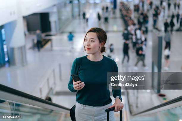 beautiful young asian woman using smartphone while riding on escalator in the airport. female tourist with mobile phone on her way to departure gate in airport. lifestyle and technology. business travel. travel and vacation concept - come & get it stock pictures, royalty-free photos & images