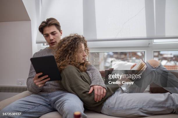 young woman reading book on cauch while her boyfriend is reading e-book - e book stockfoto's en -beelden