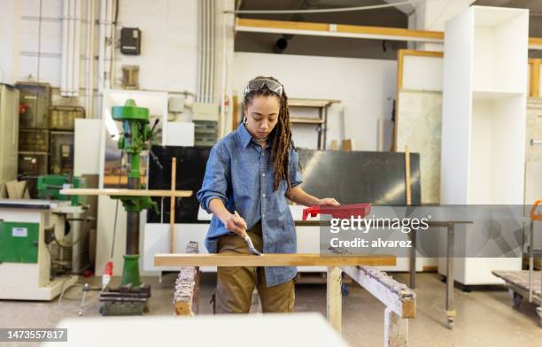 giovane donna che ricopre un pezzo di legno con vernice protettiva al laboratorio di falegnameria - wooden surface finishes foto e immagini stock