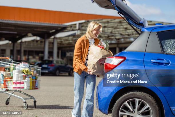 young woman loading her car with groceries after shopping - achterbak-van-auto stockfoto's en -beelden