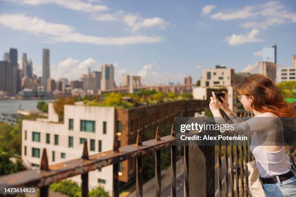 eine junge frau fotografiert an einem sonnigen sommertag die brooklyn heights promenade. - brille fluss stadt stock-fotos und bilder