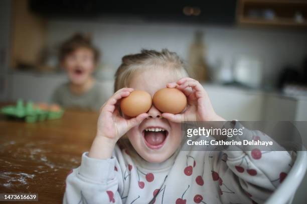 a little girl hiding her eyes with eggs as she cooking with her brother - cookery stock pictures, royalty-free photos & images