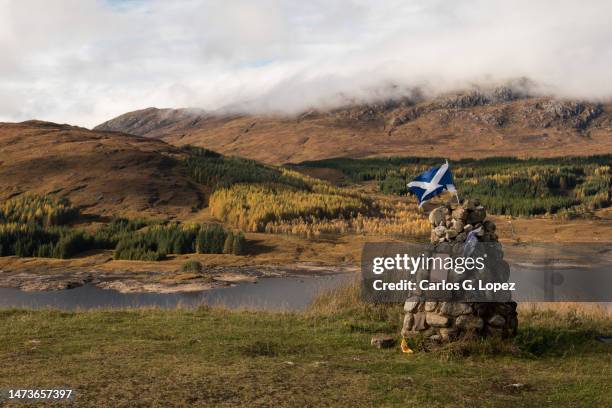 view of a cairn with the scotland flag on top with a hill partially covered with autumn colour trees on the background in a misty day in the scottish highlands, uk - glencoe schotland stockfoto's en -beelden