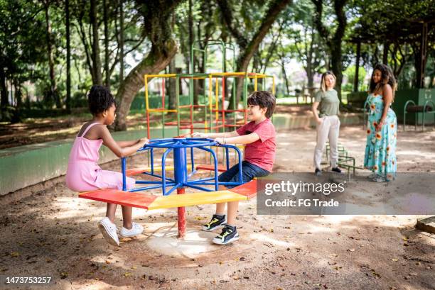 kids playing in a merry-go-round while their mothers look in a park - outdoor play equipment stock pictures, royalty-free photos & images